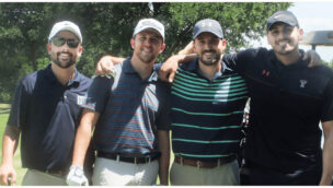 Four men in golf attire stand together on a golf course, smiling at the camera.