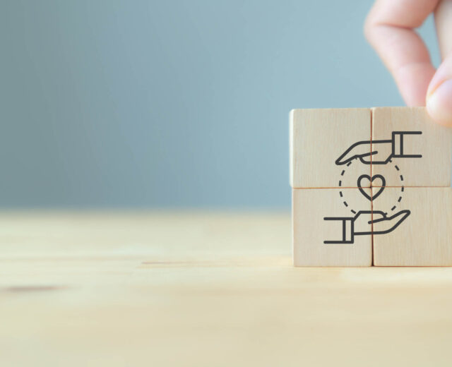 A hand places wooden blocks with a drawing of two hands surrounding a heart, symbolizing care and support, on a light wooden surface.
