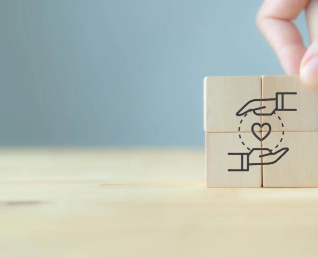 A hand places wooden blocks with a drawing of two hands surrounding a heart, symbolizing care and support, on a light wooden surface.