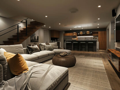 A modern basement living area with a large sectional sofa, a round ottoman, a textured rug, and a kitchen island with barstools. Wooden stairs lead to the upper floor.