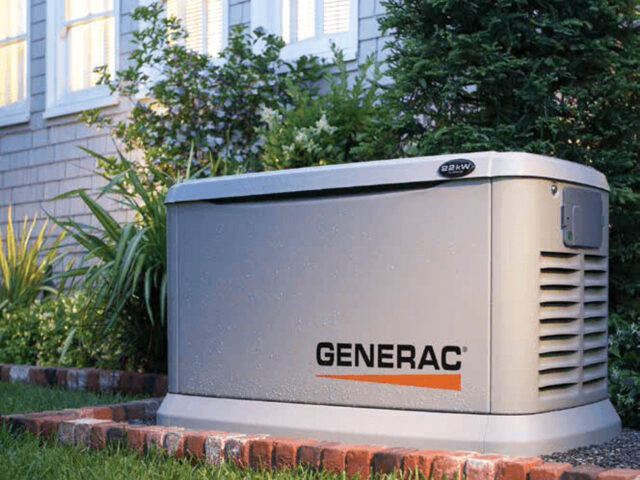 A Generac standby generator is placed next to a house, surrounded by grass and shrubs.