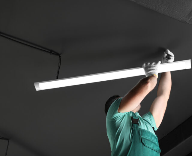 A worker in green overalls installs a fluorescent light fixture on a dark ceiling.