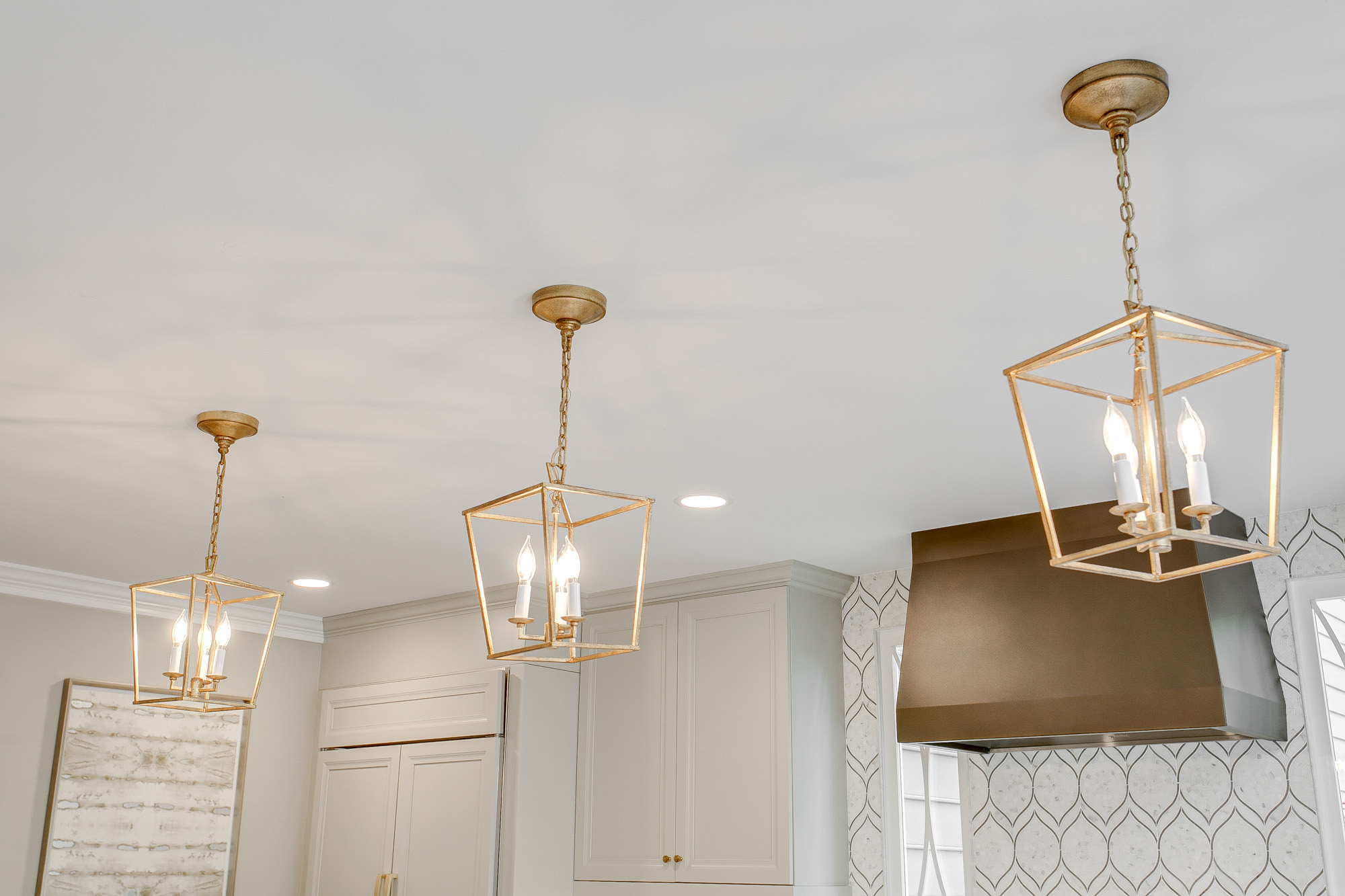 Three gold geometric pendant lights hang from the ceiling in a modern kitchen with white cabinets and a geometric backsplash.