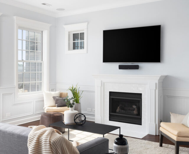 A bright living room with a gray sofa, armchairs, and a coffee table. A flat-screen TV is mounted above a white fireplace. Large windows allow natural light in.