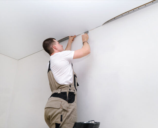 Person in overalls installing or adjusting a ceiling panel in a white room.