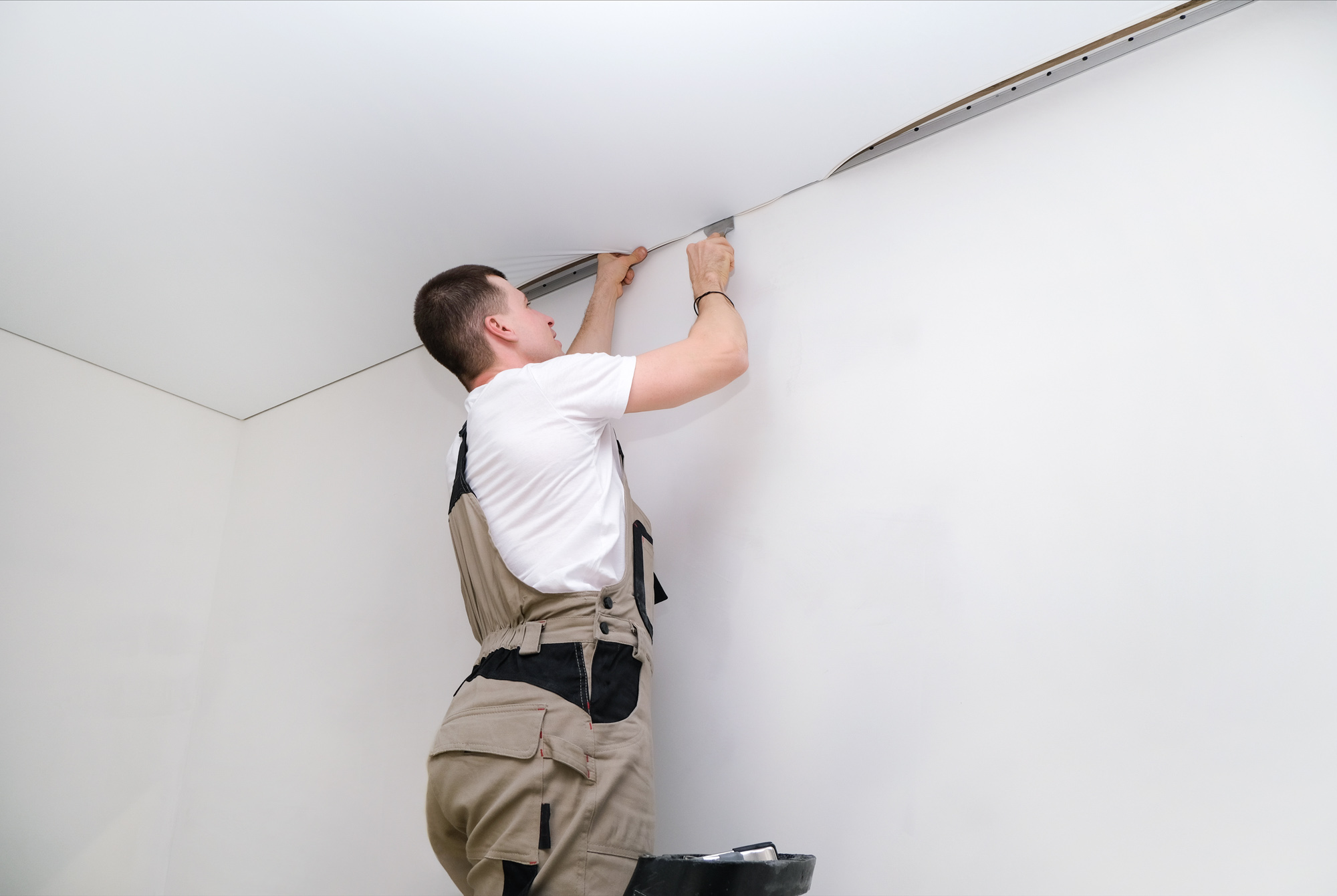 Person in overalls installing or adjusting a ceiling panel in a white room.