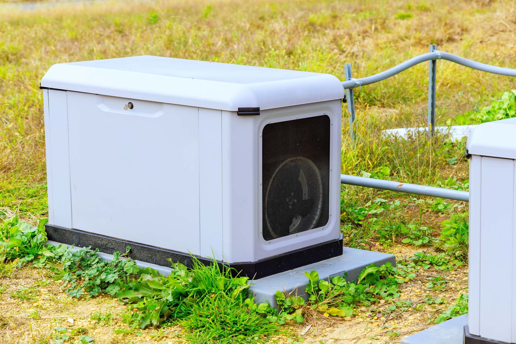 A white outdoor generator is placed on a concrete slab, surrounded by grass and some wiring.