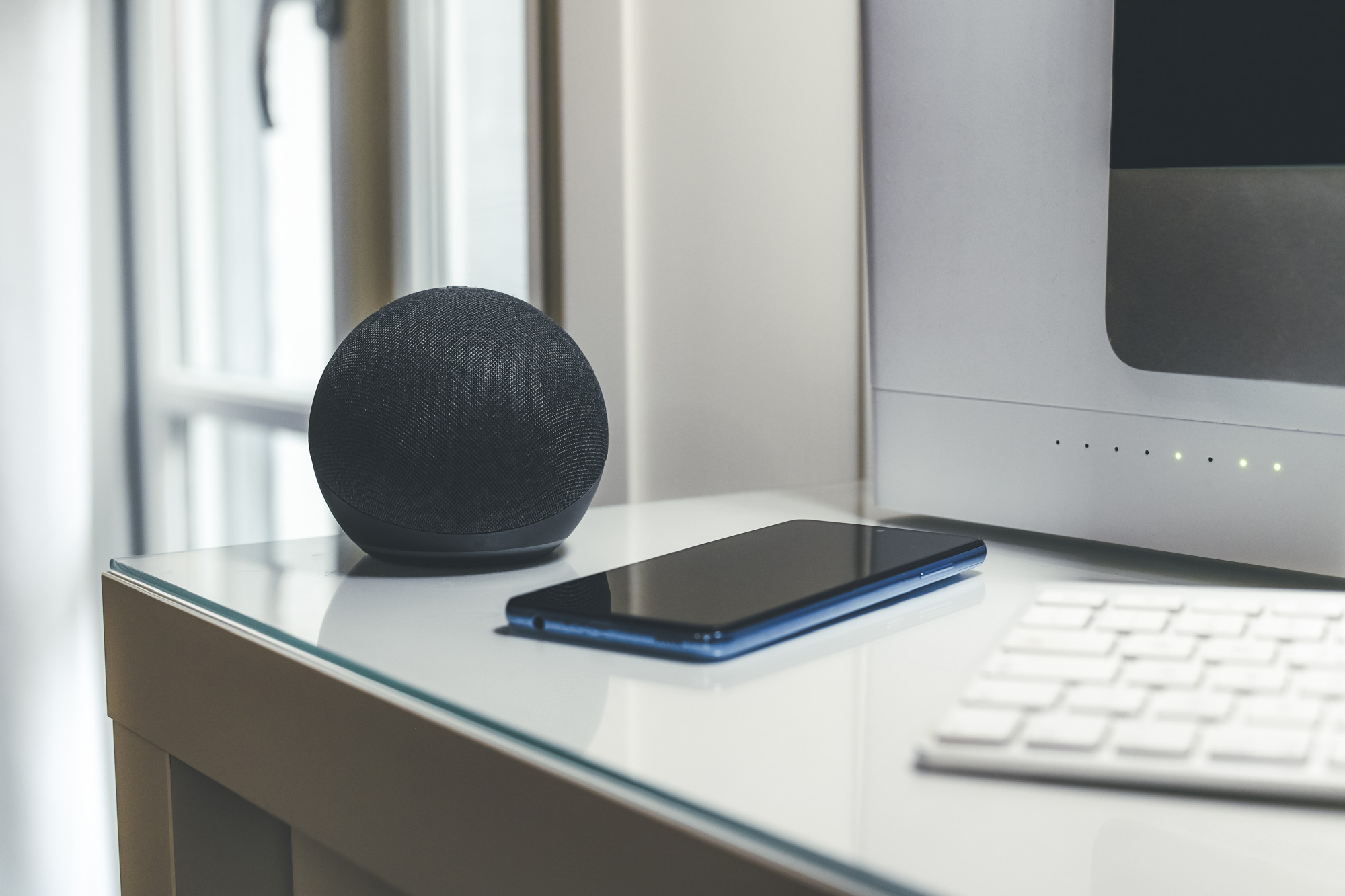 A smart speaker, smartphone, and keyboard are placed on a glass desk near a computer monitor.