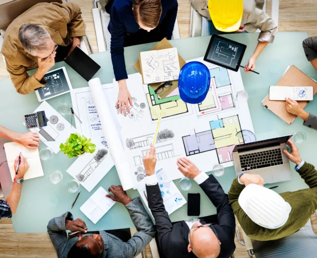 Aerial view of a diverse team wearing construction helmets and discussing blueprints at a table cluttered with papers, laptops, and tablets.