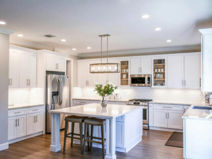 Modern kitchen with white cabinets, stainless steel appliances, and a center island with barstools. Ceiling lights and a pendant light fixture illuminate the space.