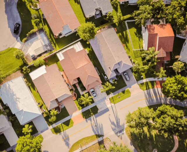 Aerial view of a suburban neighborhood with five houses, green lawns, and parked cars. The streets are curved, and the area is surrounded by trees.