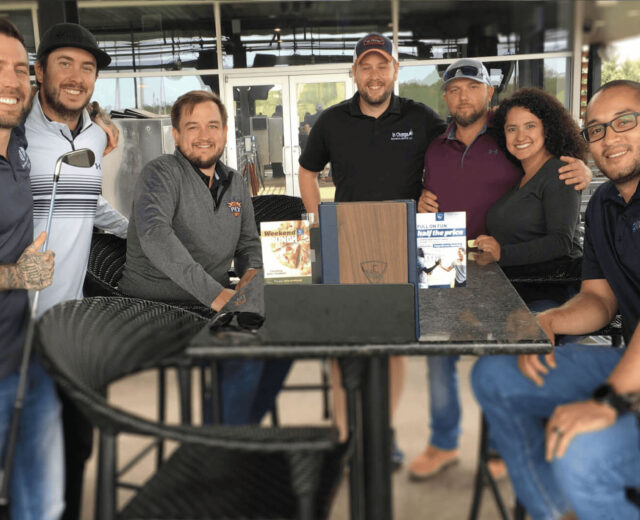 A group of seven people posing together around a high table at an outdoor patio, smiling at the camera.
