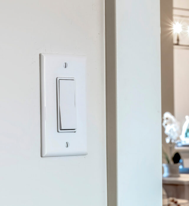 Close-up of a white light switch on a wall, with a blurred dining area and light fixture in the background.