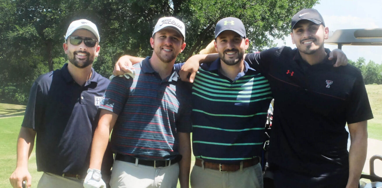 Four men in golf attire stand together on a golf course, smiling at the camera.