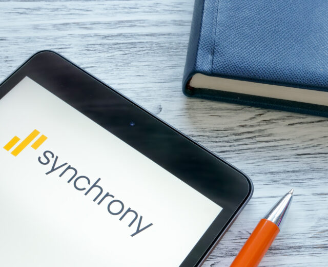Tablet displaying the Synchrony logo next to a closed blue notebook and an orange pen on a wooden surface.