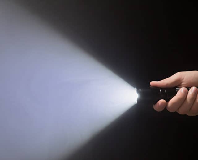 Black flashlight with a beam of light in male's hand isolated from right side of the frame on black background.