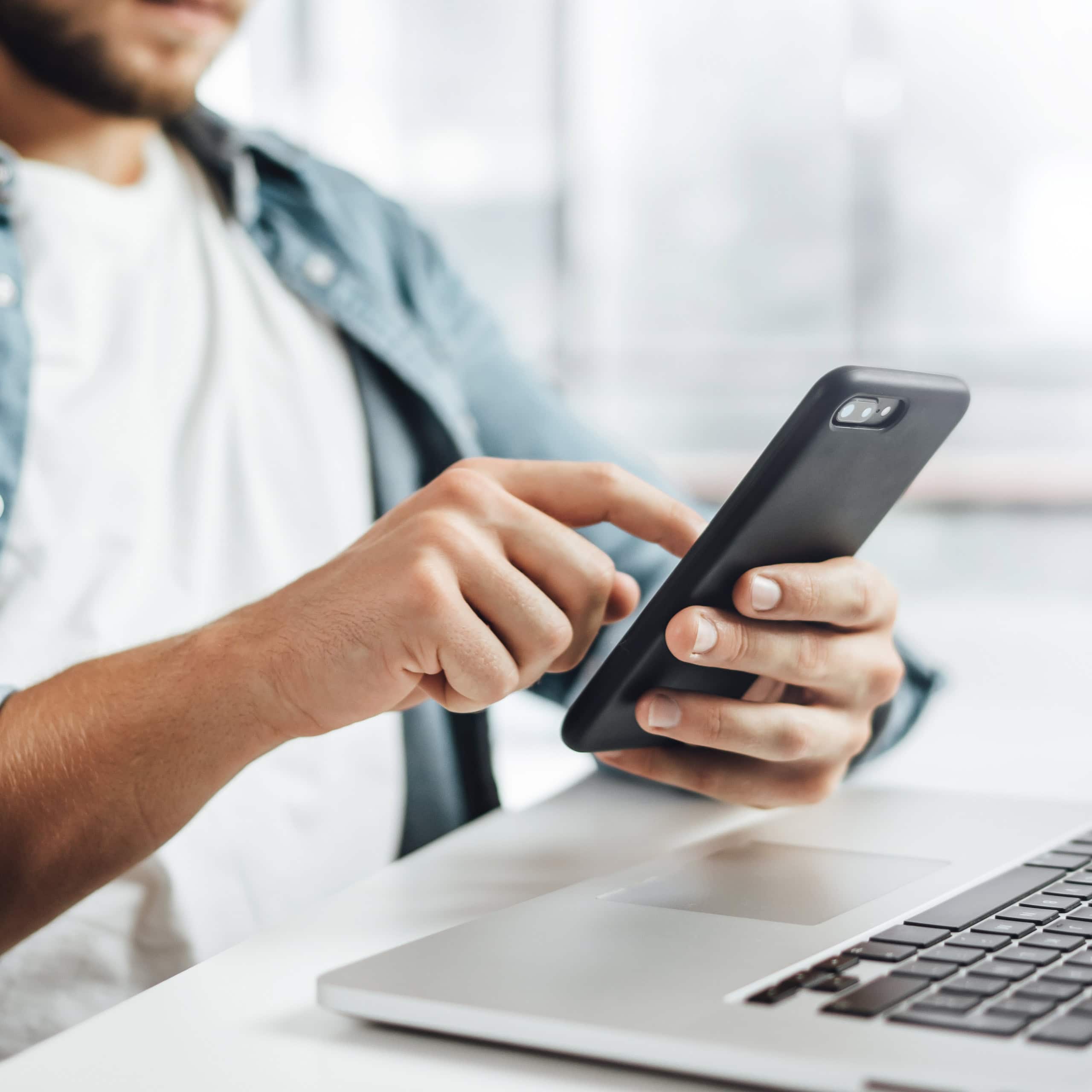 Person using a smartphone while seated at a desk with a laptop.