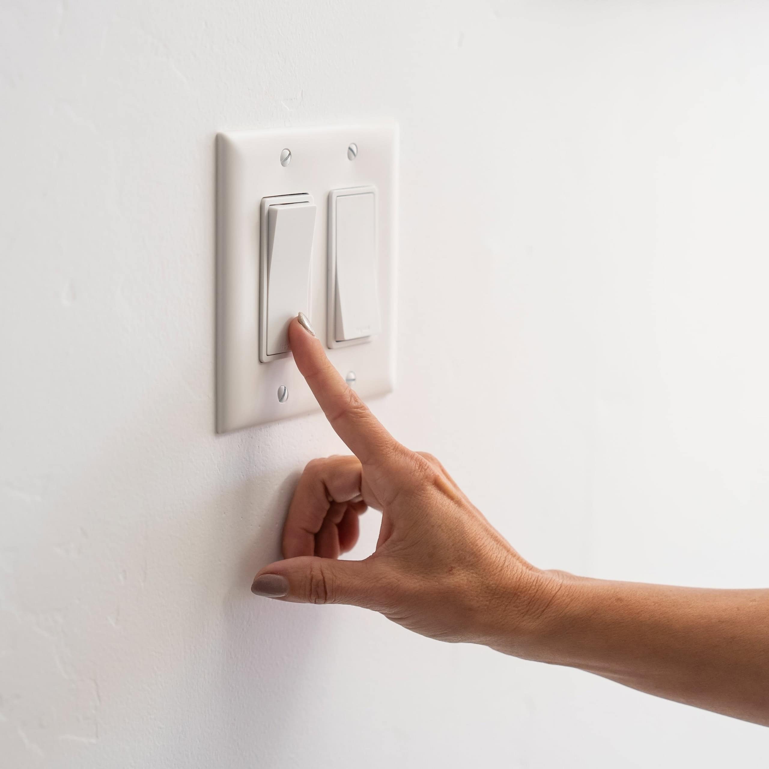 A hand reaching to flip a light switch on a white wall.